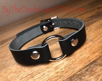 Könnte beinhalten: Schwarzer Leder-Choker mit einem silbernen Ring und zwei silbernen Druckknöpfen.