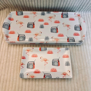 May include: A set of rectangular trays featuring a white background with a repeating pattern of pink flamingos, orange portable radios, and dark green palm leaves. The set includes one large tray and one smaller tray with raised edges.
