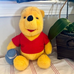 May include: A plush Winnie the Pooh bear with yellow fur, a red shirt, and a black nose. The bear is sitting upright and holding a small, round, light blue pot in its right hand. The toy has a soft, fuzzy texture and a friendly, smiling expression.
