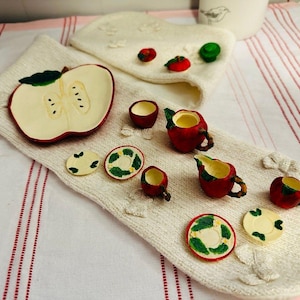 May include: A miniature tea set featuring apple-shaped pieces, including a teapot, cups, and saucers with red and green details, arranged on a cream-colored knitted fabric base with a large apple slice ornament and small decorative apple accents.