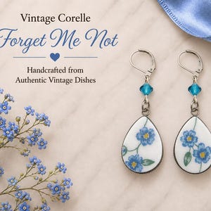 May include: A pair of teardrop-shaped earrings featuring a white ceramic base with blue forget-me-not floral patterns. Each earring includes a silver-toned lever-back hook and a small, faceted blue crystal bead accent above the ceramic pendant.