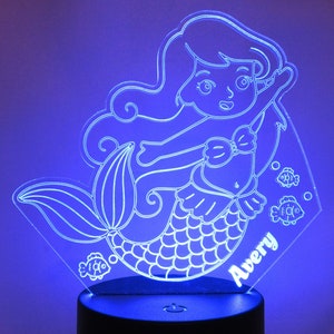 May include: A white acrylic night light shaped like a mermaid with a name "Avery" inscribed on the base. The mermaid has long flowing hair, a seashell bra, and a fishtail. There are small fish designs around the mermaid.