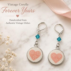 May include: A pair of dangle earrings featuring circular charms made from white ceramic with a pink heart pattern. Each earring has a silver-toned leverback hook and a small, faceted teal blue crystal bead accent above the circular charm.