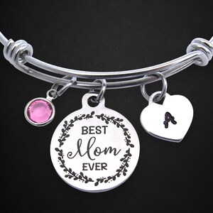 May include: Silver adjustable bracelet with three charms. One charm is a pink gemstone, another is a round pendant with "Best Mom Ever" text, and the third is a heart with the letter "A".