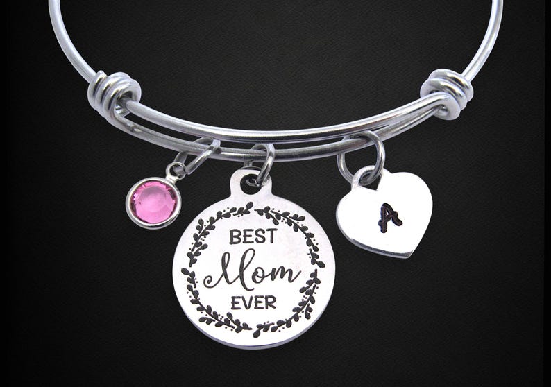 May include: Silver adjustable bracelet with three charms. One charm is a pink gemstone, another is a round pendant with "Best Mom Ever" text, and the third is a heart with the letter "A".