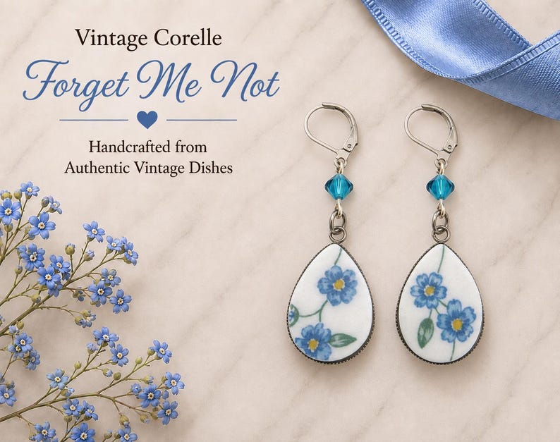 May include: A pair of teardrop-shaped earrings featuring a white ceramic base with blue forget-me-not floral patterns. Each earring includes a silver-toned lever-back hook and a small, faceted blue crystal bead accent above the ceramic pendant.