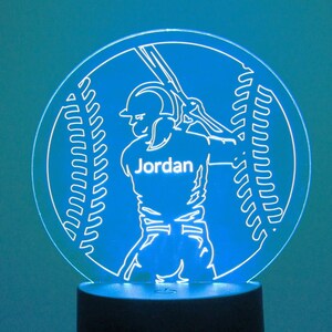 May include: White silhouette of a baseball player holding a bat with the name "Jordan" on the jersey, inside a white outline of a baseball with stitching, on a blue background. The image is a night light.
