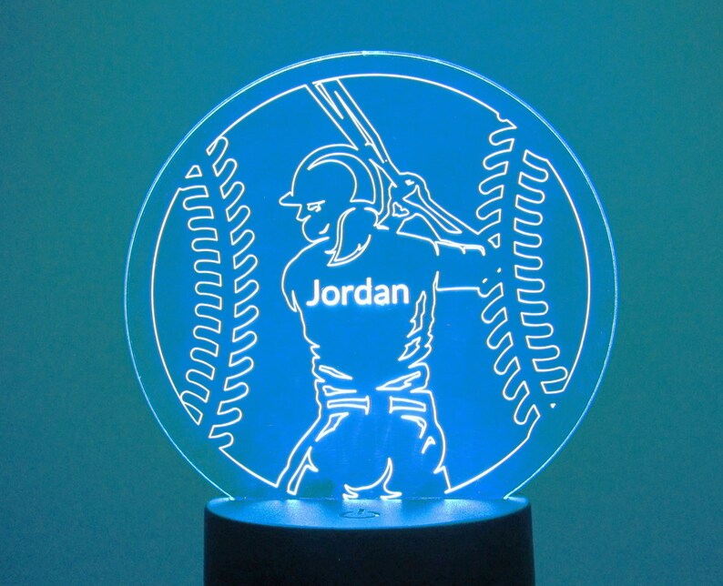 May include: White silhouette of a baseball player holding a bat with the name "Jordan" on the jersey, inside a white outline of a baseball with stitching, on a blue background. The image is a night light.