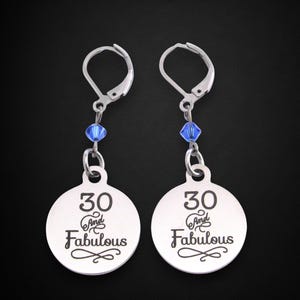May include: A pair of silver-toned earrings. Each earring features a round charm with the inscription "30 and Fabulous" in black script. A blue crystal bead hangs between the charm and the earring hook.