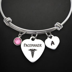 May include: Silver-toned adjustable bangle bracelet with three charms. One charm is a pink gemstone, and the other two are heart-shaped. One heart charm has the letter "A", and the other says "Pacemaker" with a medical symbol.