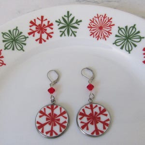 May include: A pair of festive earrings with a red snowflake design on a white background, set in a silver-colored frame. Each earring has a red bead and a lever-back closure. The earrings are displayed on a white plate with red and green snowflake patterns.
