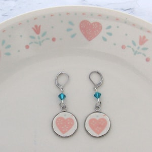 May include: A pair of silver-tone leverback earrings. Each earring features a small, round charm with a pink heart design. A blue crystal bead hangs between the charm and the leverback clasp. The earrings are displayed on a white plate with a pink heart and floral pattern.