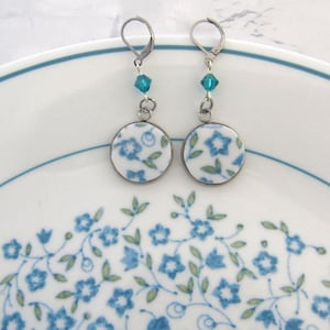 May include: A pair of dangle earrings featuring circular white charms with a blue and green floral pattern. Each earring is suspended from a silver-toned leverback hook and accented with a single small, faceted turquoise-colored crystal bead.