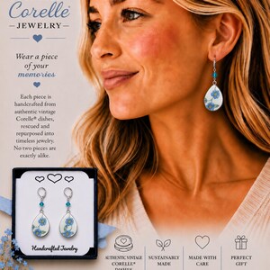 May include: Dangle earrings featuring teardrop-shaped white ceramic pendants with a blue floral pattern. Each earring includes a small blue crystal bead on a silver-toned metal hook. The earrings are displayed in a black box with white text.