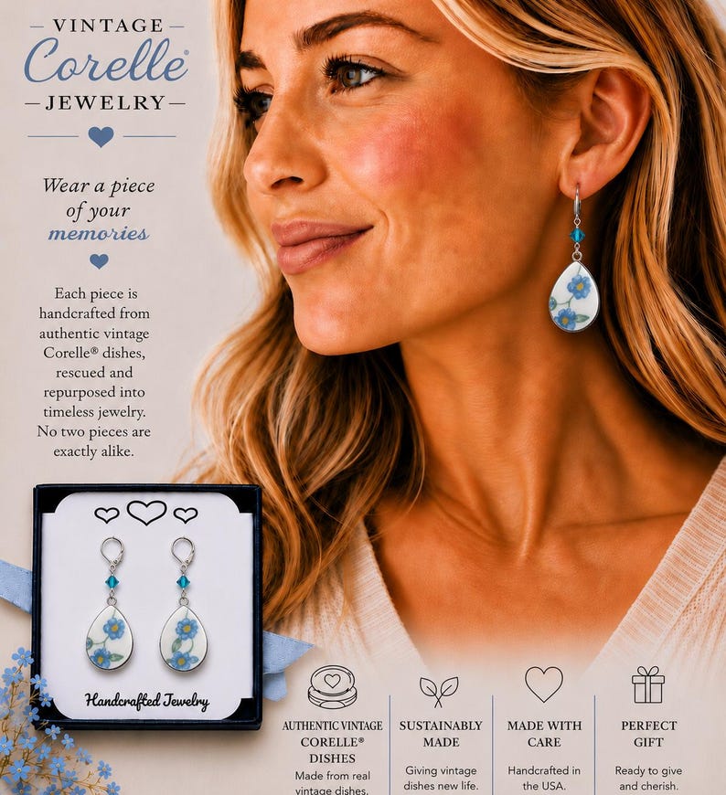 May include: Dangle earrings featuring teardrop-shaped white ceramic pendants with a blue floral pattern. Each earring includes a small blue crystal bead on a silver-toned metal hook. The earrings are displayed in a black box with white text.