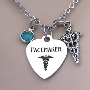 May include: Silver-toned necklace with a heart-shaped pendant that reads "Pacemaker" and features a medical symbol. A teal-colored gemstone and a medical symbol charm are also attached to the chain. The necklace is a medical-themed accessory.