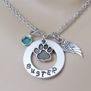 May include: Silver necklace with a round charm that says "Buster" and has a paw print charm inside the circle. The necklace also has a small teal blue crystal charm and a silver angel wing charm.