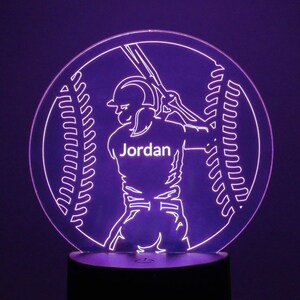 May include: A purple LED night light with a baseball player silhouette holding a bat. The player is wearing a jersey with the name "Jordan" on the front. The background is a baseball with stitching.