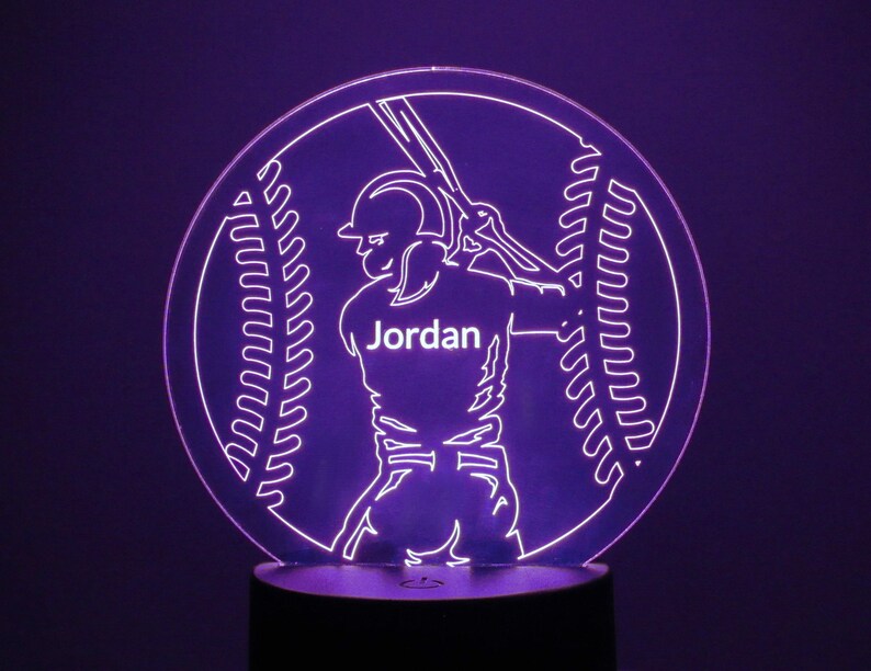 May include: A purple LED night light with a baseball player silhouette holding a bat. The player is wearing a jersey with the name "Jordan" on the front. The background is a baseball with stitching.