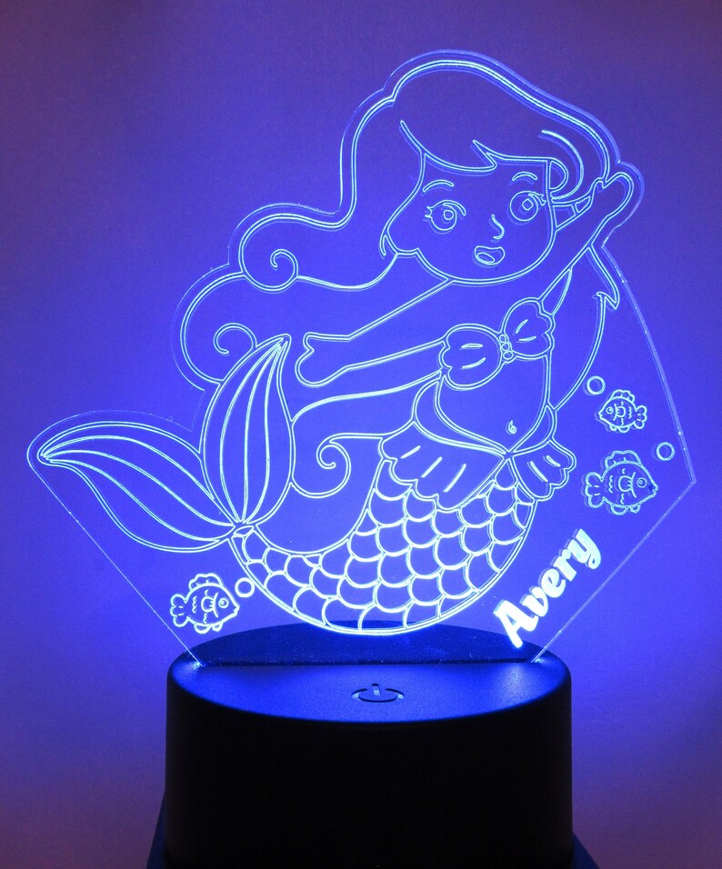 May include: A white acrylic night light shaped like a mermaid with a name "Avery" inscribed on the base. The mermaid has long flowing hair, a seashell bra, and a fishtail. There are small fish designs around the mermaid.