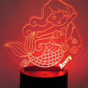 May include: A red LED night light shaped like a mermaid with a tail, scales, and a bow. The mermaid is holding a fish in her hand. The night light has the name "Avery" written on it.