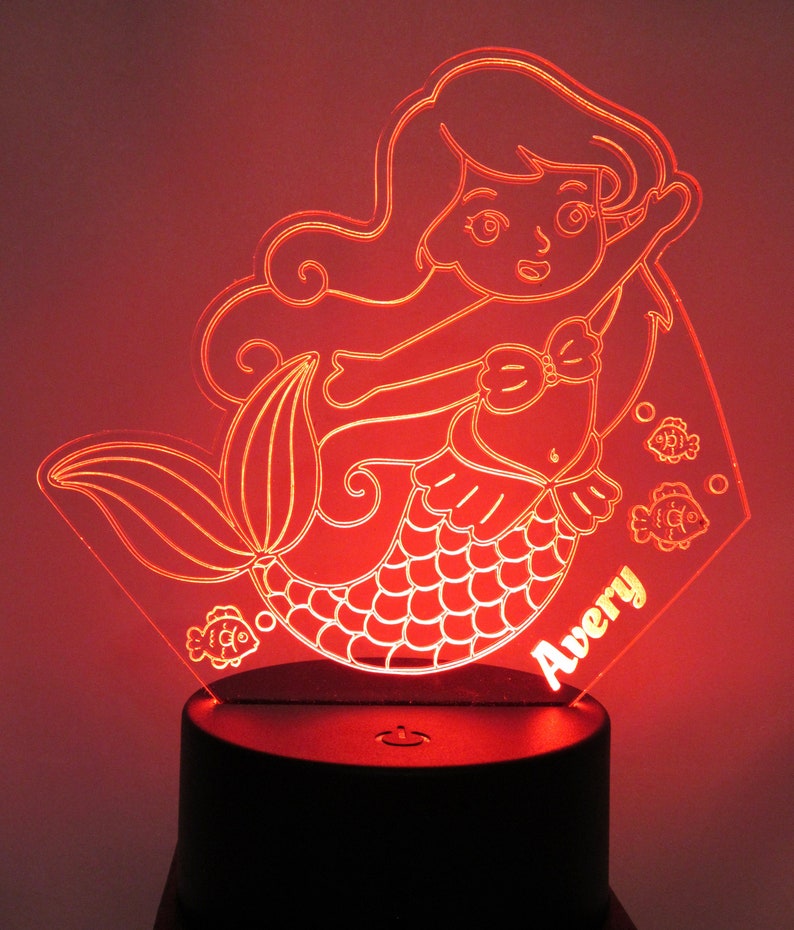 May include: A red LED night light shaped like a mermaid with a tail, scales, and a bow. The mermaid is holding a fish in her hand. The night light has the name "Avery" written on it.