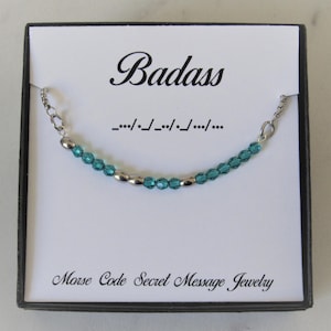 May include: A teal blue and silver beaded necklace with the word "Badass" and its Morse code translation, "--/./--/./---/.", printed on a white card in a black box. The card also reads "Morse Code Secret Message Jewelry".