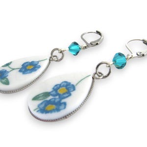 May include: A pair of drop earrings featuring white teardrop-shaped pendants with a blue and yellow floral pattern. Each earring includes a faceted turquoise crystal bead connected to a silver-toned metal leverback closure for secure attachment.