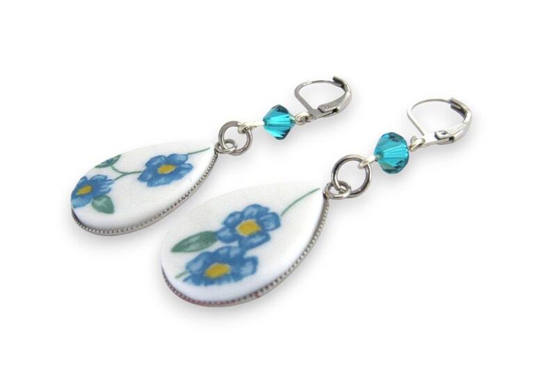 May include: A pair of drop earrings featuring white teardrop-shaped pendants with a blue and yellow floral pattern. Each earring includes a faceted turquoise crystal bead connected to a silver-toned metal leverback closure for secure attachment.