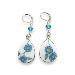May include: A pair of drop earrings featuring white, teardrop-shaped ceramic pendants decorated with blue flowers, green leaves, and yellow centers. Each earring includes a single faceted turquoise crystal bead and a silver-toned leverback clasp.