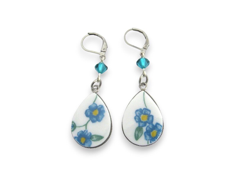 May include: A pair of drop earrings featuring white, teardrop-shaped ceramic pendants decorated with blue flowers, green leaves, and yellow centers. Each earring includes a single faceted turquoise crystal bead and a silver-toned leverback clasp.