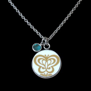 May include: A silver chain necklace featuring a round pendant with a white background and a gold butterfly graphic. A small, circular, teal-colored gemstone charm hangs next to the main pendant, both suspended from a simple silver link chain.
