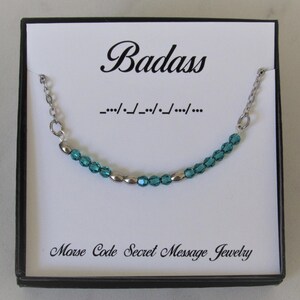 May include: A teal blue and silver beaded necklace with a silver chain. The necklace is in a white box with the word "Badass" written in black cursive and the morse code for "Badass" written below it. The box also says "Morse Code Secret Message Jewelry".