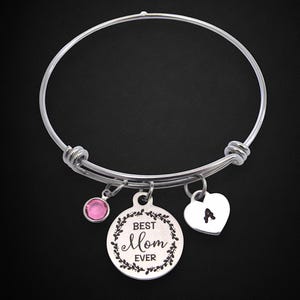 May include: Silver-toned bangle bracelet with three charms. One charm is a pink gemstone, another is a round pendant with the text "Best Mom Ever" and a floral border, and the third is a heart-shaped charm with the letter "A".