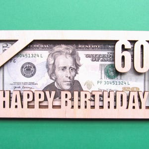 May include: A wooden card with a cutout for a dollar bill. The bill is visible through the cutout and the number "60" is cut out of the wood above the bill. The words "Happy Birthday" are cut out of the wood below the bill.
