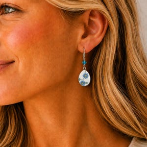 May include: A teardrop-shaped earring featuring a white ceramic base with a blue floral pattern. The earring is suspended from a silver-toned hook and accented with a small, faceted, bright blue crystal bead positioned above the ceramic pendant.