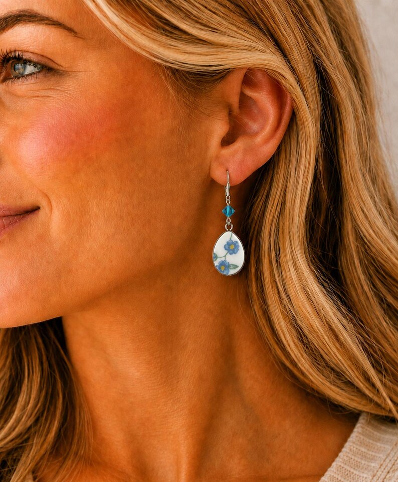 May include: A teardrop-shaped earring featuring a white ceramic base with a blue floral pattern. The earring is suspended from a silver-toned hook and accented with a small, faceted, bright blue crystal bead positioned above the ceramic pendant.