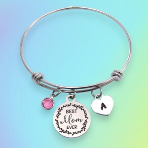 May include: A silver-colored bangle bracelet with three charms. One charm is a pink gemstone, another is a heart with the letter "A", and the third is a round charm with the words "Best Mom Ever" inside a floral wreath.