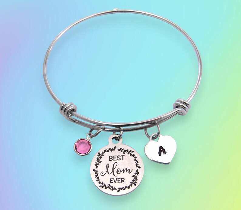 May include: A silver-colored bangle bracelet with three charms. One charm is a pink gemstone, another is a heart with the letter "A", and the third is a round charm with the words "Best Mom Ever" inside a floral wreath.