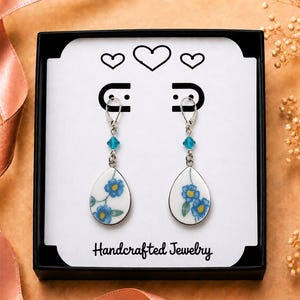 May include: A pair of drop earrings featuring white teardrop-shaped charms with blue floral patterns, accented by small turquoise beads and silver-toned lever-back hooks, presented in a black box with the text Handcrafted Jewelry.