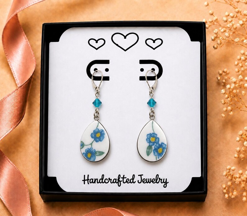 May include: A pair of drop earrings featuring white teardrop-shaped charms with blue floral patterns, accented by small turquoise beads and silver-toned lever-back hooks, presented in a black box with the text Handcrafted Jewelry.