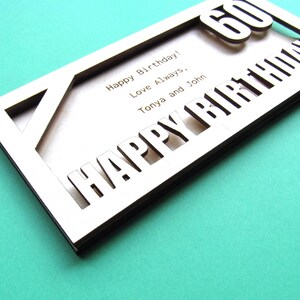 May include: A wooden card with the words "Happy Birthday" cut out in the front. The number "60" is visible through the cut-out letters. The card also has the text "Happy Birthday! Love Always, Tonya and John" written on the inside.