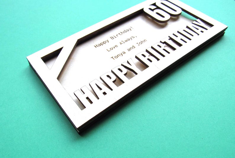 May include: A wooden card with the words "Happy Birthday" cut out in the front. The number "60" is visible through the cut-out letters. The card also has the text "Happy Birthday! Love Always, Tonya and John" written on the inside.