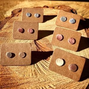 May include: A collection of handcrafted stud earrings displayed on small, brown cardboard cards. The earrings are in pairs, with silver and copper-toned metal options. The earrings have a hammered texture and a simple, round design.