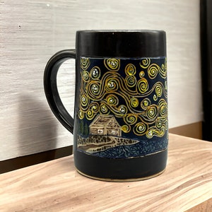 May include: Black ceramic mug with a hand-painted design of a starry night sky with a small house in the foreground. The design is in shades of yellow, white, and blue.