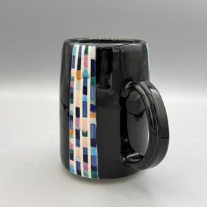 Pode incluir: Caneca de cerâmica preta com acabamento brilhante. Uma faixa vertical de retângulos coloridos em rosa, azul, verde e branco adorna a lateral. A caneca tem uma alça curva e uma base lisa e arredondada.