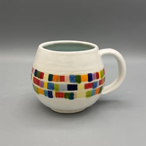 Pode incluir: Uma caneca de cerâmica branca com corpo arredondado e uma alça. A caneca apresenta uma faixa de padrões retangulares coloridos em vermelho, amarelo, azul, verde e laranja. O interior da caneca é azul claro.