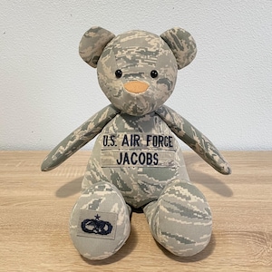 May include: A stuffed teddy bear crafted from grey and light grey digital camouflage fabric. The bear features black button eyes, a tan nose, and a chest patch embroidered with U.S. AIR FORCE and JACOBS. A dark blue military insignia is on one foot.