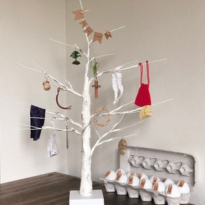May include: A white birch tree with a white trunk and branches decorated with ornaments for an Advent calendar. The ornaments include a small potted tree, a crown of thorns, a red bag, a wooden cross, a small gourd, a piece of fabric, and a small white bag. The tree is standing on a white base and is surrounded by 12 white eggs with numbers on them.