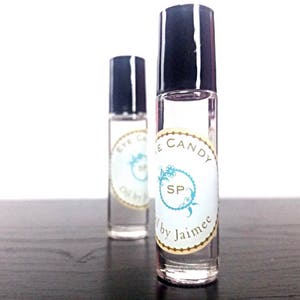 May include: Two clear glass roller bottles with black caps and labels. The label on the front bottle says "Eye Candy" and the label on the back bottle says "Eye Candy SP by Jaimee".